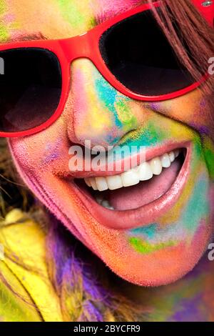 Felice faccia emozionata della donna coperta di polvere di colore arcobaleno. Giovane donna allegra in occhiali da sole e con viso ricoperto di pigmento brillante multicolore Foto Stock