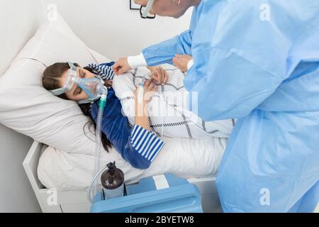 Il medico controlla il collegamento del ventilatore. L'adolescente si trova su un letto d'ospedale. Maschera di ossigeno sul tratto respiratorio. Foto Stock