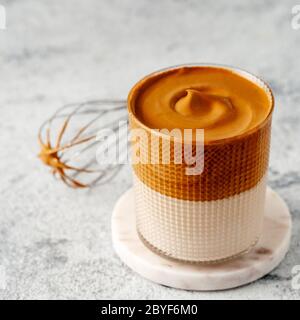 Bevanda coreana alla moda - caffè dalgona. Vaso in vetro con bevanda fredda o calda - caffè istantaneo montato con latte e frusta su fondo grigio in cemento con spazio per la copia di testo o design Foto Stock