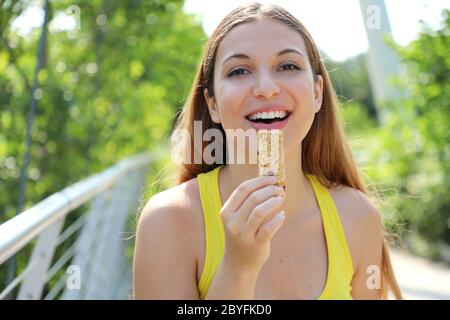 Donna del fitness che mangia una barra energetica guardando la macchina fotografica. Primo piano di ragazza sportiva che mangia uno spuntino di cereali all'aperto. Foto Stock