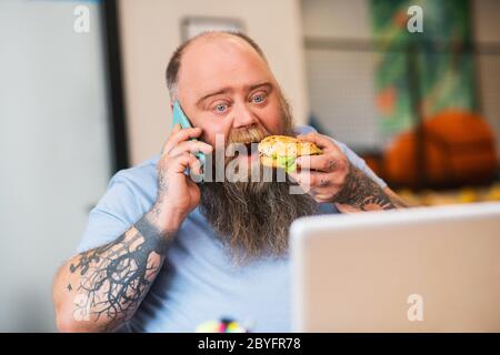 Uomo calvo che parla al telefono e che mangia un hamburger Foto Stock