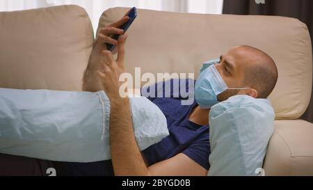 Uomo con maschera di protezione durante il blocco del coronavirus scorrimento sullo smartphone mentre abbracciano un cuscino. Foto Stock