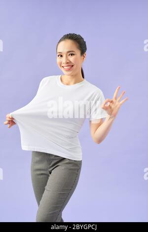 Donna giovane stupita in t-shirt bianca che mostra i suoi risultati di dieta su sfondo viola Foto Stock