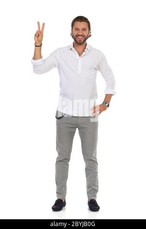 Elegante bell'uomo in mocassini, pantaloni grigi e camicia bianca è in piedi, mostrando il segno della mano di pace e sorridendo. Studio di lunghezza intera isolato su Foto Stock