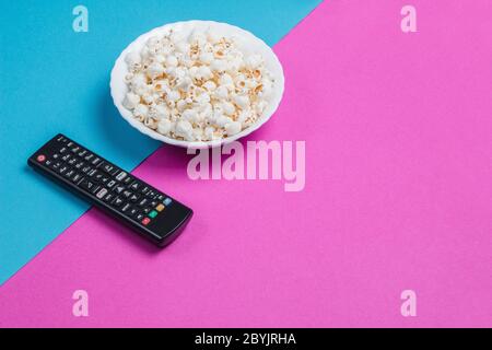 Ciotola bianca di popcorn su sfondo blu e viola con telecomando TV nero Foto Stock