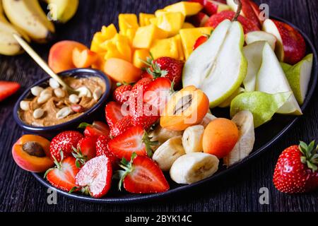 Piatto di frutta arcobaleno di frutti freschi e bacche con salsa di arachidi: Fragole, mango tropicale, banana, mele, pere, albicocche e arachidi su un blac Foto Stock