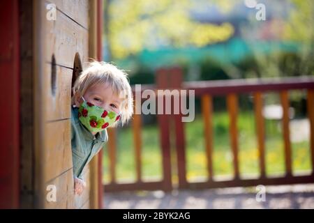 Bambino biondo piccolo toddler che indossa una maschera protettiva, giocando sul parco giochi, primavera soleggiato giorno Foto Stock