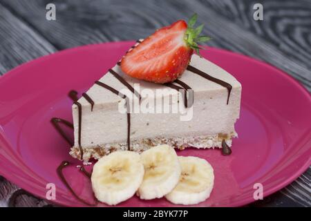 Cheesecake su un piatto. Guarnito con fragole, banane e cioccolato. Su tavole di pino spazzolato verniciate in bianco e nero. Foto Stock
