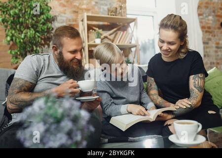 Famiglia felice a casa passare il tempo insieme. Divertendosi, sembra allegro e bello. Libro di lettura di madre, padre e figlio durante gli studi remoti della scuola. Aiuto dei genitori. Infanzia, concetto di vita domestica. Foto Stock