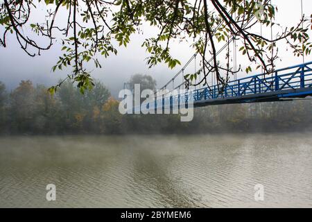 Ponte sospeso sul fiume sola, nebbia mattina d'autunno. Paesaggio mistico Foto Stock