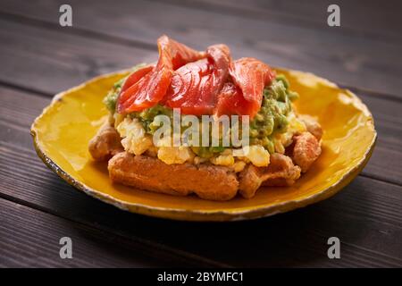 Waffle belgi con uova strapazzate, avocado e salmone su tavola di legno. Piatto, pancetta. Variazione colazione keto Foto Stock