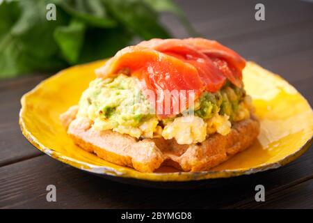 Waffle belgi con uova strapazzate, avocado e salmone su tavola di legno. Piatto, pancetta. Variazione colazione keto Foto Stock