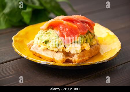 Waffle belgi con uova strapazzate, avocado e salmone su tavola di legno. Piatto, pancetta. Variazione colazione keto Foto Stock