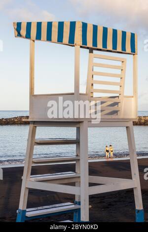 Donna più giovane che aiuta una donna anziana nel mare a Playa Beril incorniciato attraverso le gambe del posto di osservazione bagnino, Costa Adeje, Tenerife, Canary i Foto Stock