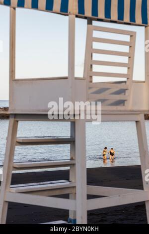 Donna più giovane che aiuta una donna anziana nel mare a Playa Beril incorniciato attraverso le gambe del posto di osservazione bagnino, Costa Adeje, Tenerife, Canary i Foto Stock