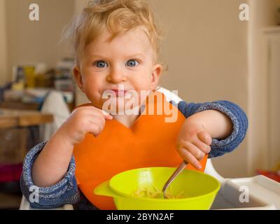 Bambino di un anno con espressione divertente mangiare con posate a casa Foto Stock