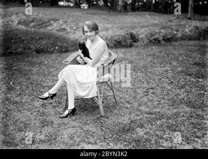 Inizio del XX secolo Inglese vintage fotografia in bianco e nero che mostra una giovane donna, seduta su una sedia in vimini, in un giardino, che tiene un gatto. La donna mostra moda e stile capelli del periodo, 1920. Foto Stock