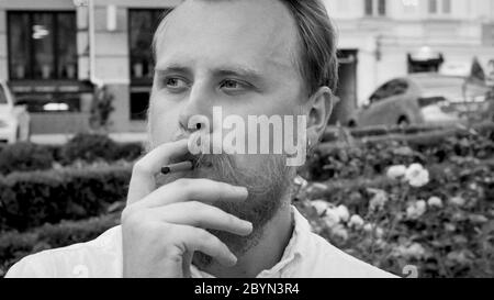 Ritratto in bianco e nero di giovane elegante con barba fumando sigaretta nel parco Foto Stock