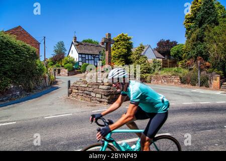 Ciclista che attraversa il pittoresco villaggio di campagna di Eaton Tarporley Cheshire, un villaggio di campagna, con una croce di pietra nel centro del paese Foto Stock