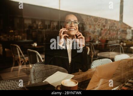 Donna che usa un telefono cellulare in un bar. Donna seduta all'interno della caffetteria che parla al telefono con un computer portatile e una tazza di caffè sul tavolo. Foto Stock