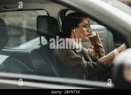 Donna d'affari con auricolari che guidano la sua automobile all'ufficio. Donna in abbigliamento da formalwear che guida al lavoro. Foto Stock
