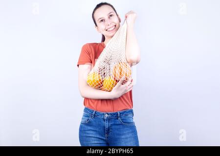 La giovane ragazza preme borsa a corda con arance e sorrisi, su sfondo grigio Foto Stock