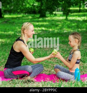 Giovane donna e bambina in abbigliamento sportivo con una mela verde e seduta sul tappeto in parco. Foto Stock