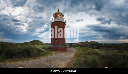 Faro di mattoni rossi, di sera, sotto un cielo scuro, a Kampen, sulla riserva naturale dell'isola di Sylt, in Germania. Faro illuminato su dune erbose panorama. Foto Stock