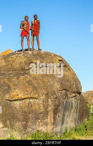 Due giovani guerrieri Masai stanno in piedi su una grande pietra in abiti tradizionali con una lancia contro un cielo blu. Tanzania, Africa orientale, 12 agosto 2018. Foto Stock