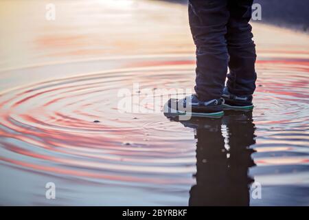 Bambino dolce, ragazzo carino con impermeabile rosso, giocando in puddle al tramonto Foto Stock