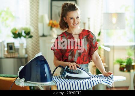 Ritratto di donna felice trendy in blusa rossa e gonna a matita grigia stiratura con generatore di vapore su asse da stiro nella casa moderna in giornata di sole. Foto Stock