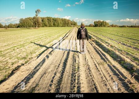 Un uomo che cammina lungo una strada sterrata tra campi, orizzonte e cielo blu, vista in giornata di sole Foto Stock