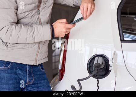 Uomo che carica un'auto elettrica presso la stazione di ricarica pubblica e paga utilizzando il suo smartphone. Concetto di emissione zero Foto Stock