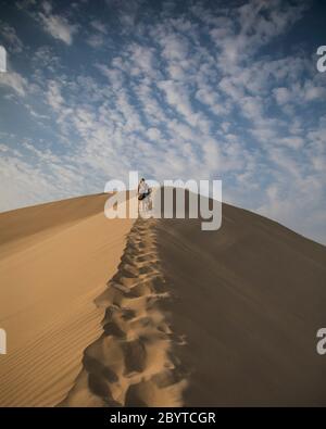 donna che arrampica una duna nel deserto, nuvole e cielo blu Foto Stock