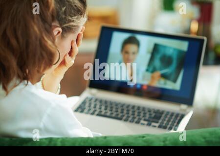 Visto da dietro donna che parla con medico utilizzando tele tecnologia di salute su un computer portatile, mentre si siede su un divano in casa in una giornata di sole. Foto Stock