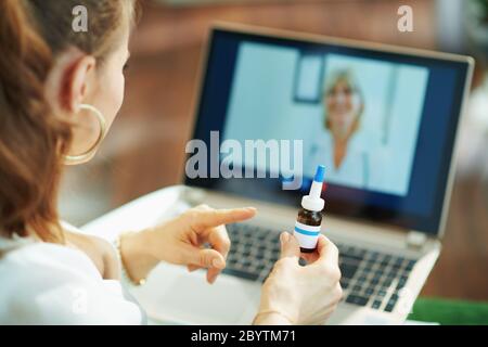 Visto da dietro la donna che mostra flacone di medicina mentre si parla con il medico utilizzando la tecnologia tele salute su un computer portatile in soggiorno in giornata di sole. Foto Stock