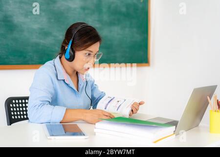 Insegnante di donna asiatica che legge un libro alto allo studente tramite video conferenza e-learning in un laptop con lavagna in classe. Scelta della posizione e distanza Foto Stock