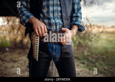 Cowboy in jeans e abbigliamento in pelle con la sua mano sul revolver, texas ranch sullo sfondo, occidentale. Vintage persona di sesso maschile con la pistola, wild west lifesty Foto Stock