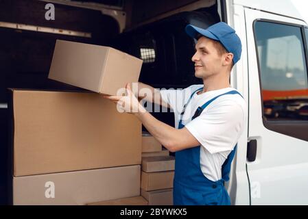 Sorridente deliveryman con scatola di cartone in auto, servizio di consegna. Uomo in confezione di cartone uniforme, spedizione maschile, lavoro di corriere Foto Stock