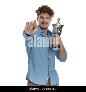 felice ragazzo in camicia denim punta dito, tenendo trofeo e sorridente, isolato su sfondo bianco, ritratto Foto Stock