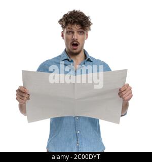 uomo casual sorpreso in denim camicia lettura giornale, in piedi isolato su sfondo bianco, ritratto Foto Stock