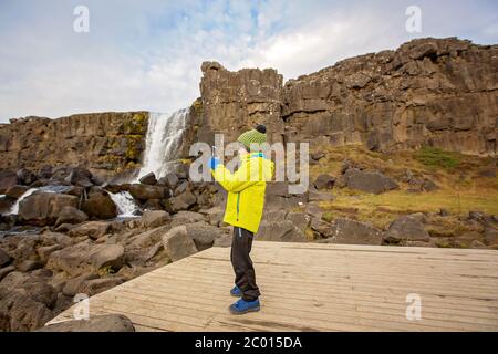 Bambino carino, ragazzo, godendo di una giornata di sole nella valle del rift del Parco Nazionale di Oxarfoss, scattando foto con il cellulare, autunno Islanda Foto Stock