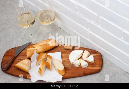 Fette di camembert, baguette e due bicchieri di vino bianco su tavola di legno. Muro bianco di mattoni come sfondo. Foto Stock