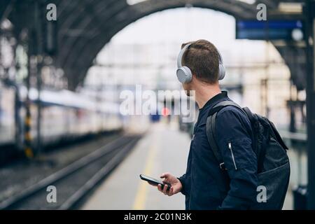 Giovane uomo con cuffie che ascolta musica e attende il treno alla stazione ferroviaria. Foto Stock