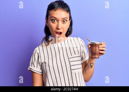 Giovane donna bevendo l'infusione di compagno spaventato e stupito con bocca aperta per sorpresa, faccia incredula Foto Stock