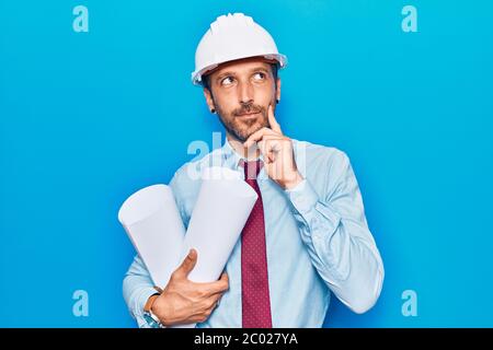 Giovane bell'uomo che indossa l'architetto hardHat che tiene i blueprints faccia seria che pensa alla domanda con la mano sul mento, pensava a confondere ide Foto Stock