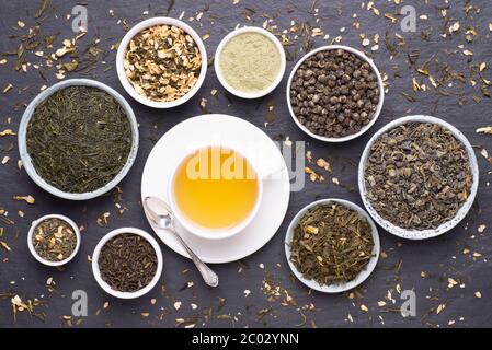 Tazza di tè verde e ciotole di varie foglie di tè secco su sfondo scuro in pietra, vista dall'alto Foto Stock