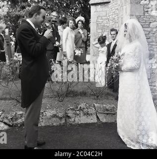 1967, storico, un ospite di matrimonio maschile nel suo vestito e code in piedi utilizzando una macchina fotografica portatile in cine per filmare la sposa nel suo vestito da matrimonio e viel fuori della chiesa, guardato da altri ospiti, Inghilterra, Regno Unito. Realizzare filmati amatoriali utilizzando piccole telecamere cine da 8 mm è stato un hobby popolare in questa epoca e i matrimoni sono sempre stati un evento da girare come un record visivo di una sposa speciale giorno. Foto Stock