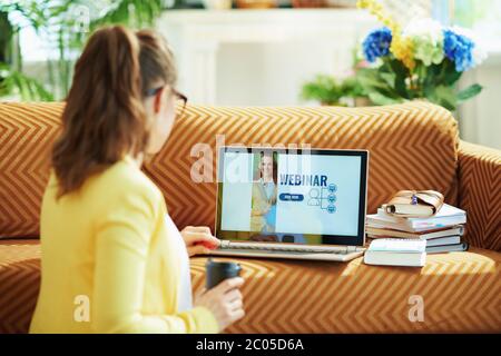 Visto da dietro donna in giacca gialla nel salotto in giornata di sole che va guardare il webinar su un computer portatile. Foto Stock