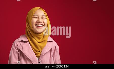 Ritratto di giovane ragazza teenage musulmana felice e fiduciosa che indossa hijab guardando la macchina fotografica e sorridendo allegro, in piedi contro sfondo rosso Foto Stock